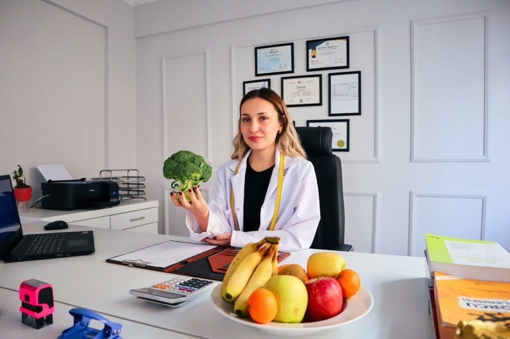 Female doctor at desk holding broccoli with fruits on desk